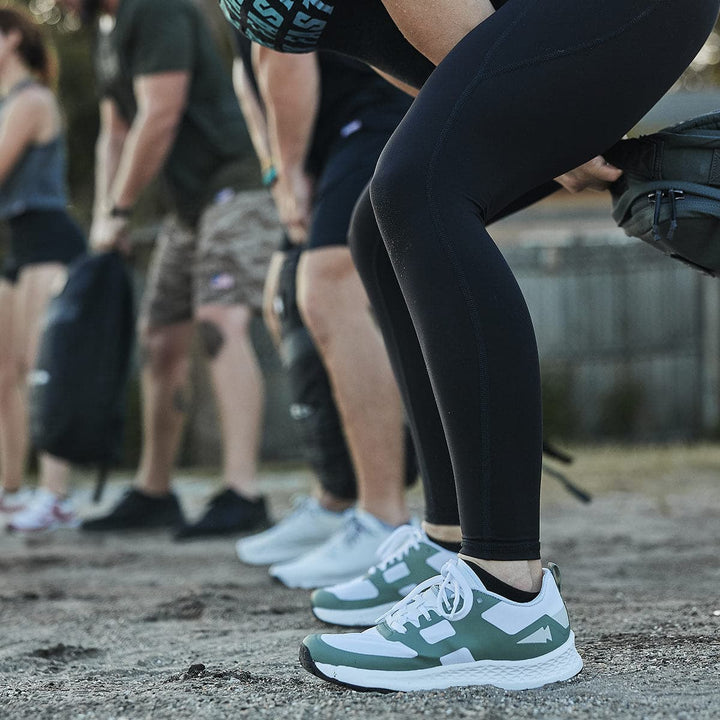 Athletic individuals are dressed in sports gear and sneakers, seemingly engaged in weightlifting on a dirt surface outdoors. In the foreground, one person's legs and their Men's Rough Runner - Seaspray shoes from GORUCK stand out, reminiscent of someone training for a challenging race.