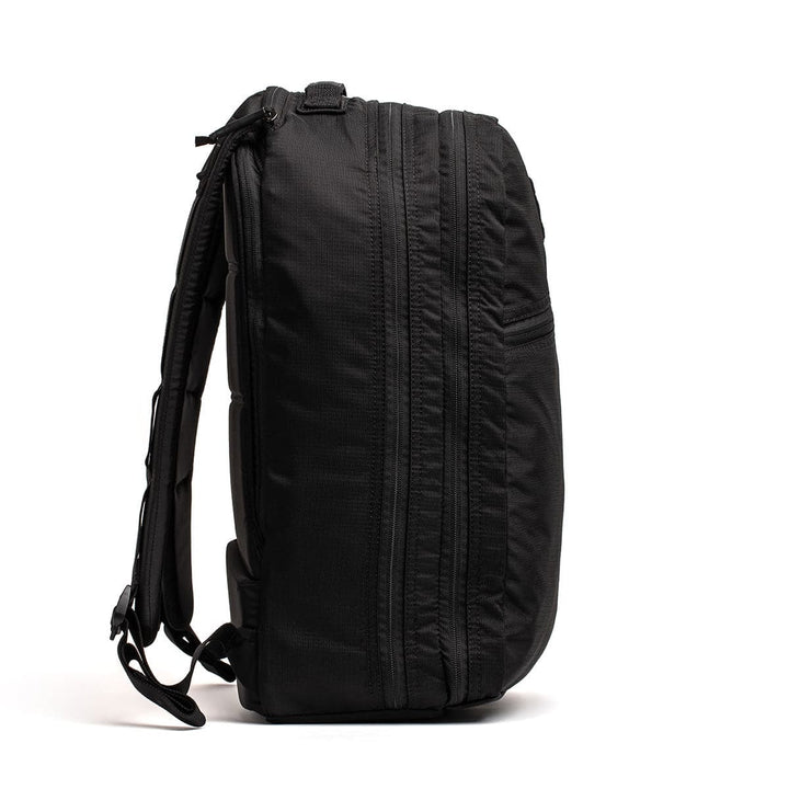 Side view of a black GORUCK Bullet Ruck Double Compartment made from ripstop nylon, featuring multiple zippers and padded shoulder straps, against a white background.