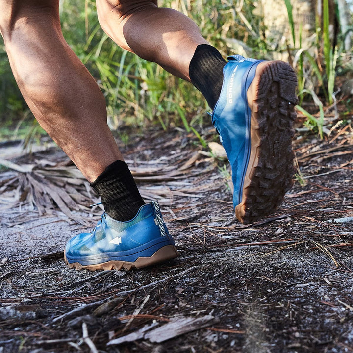 A person running on a trail in GORUCK's Mackall shoes in Legion Blue + Gum, featuring an aggressive triple compound outsole, surrounded by grass and trees.