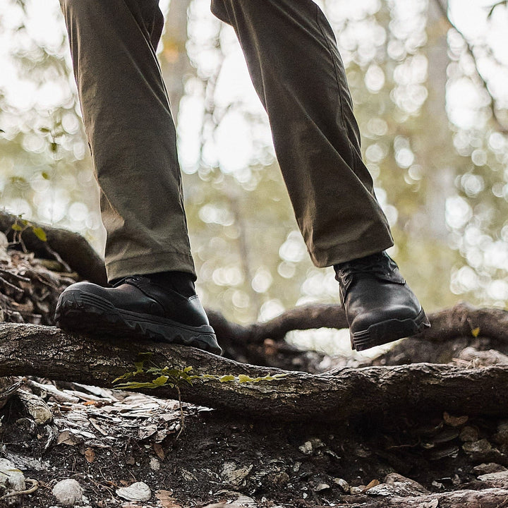 A person wearing khaki pants and GORUCK's MACV-2 Mid Top boots in black, which feature a triple compound outsole, is climbing over tree roots on a forest trail. The background is blurred, with sunlight filtering through the trees.