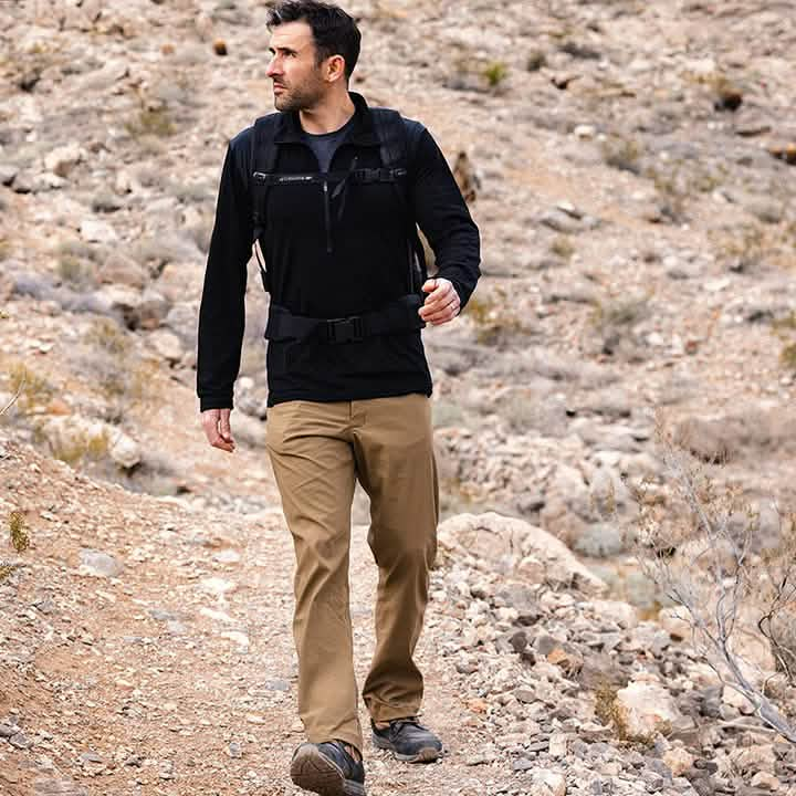 A man wearing black and khaki GORUCK’s Men’s Simple Pants - Midweight ToughDry® is hiking on a rocky trail. He carries a backpack, observing his surroundings in a dry landscape with sparse vegetation.