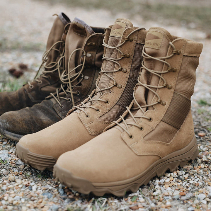 Two pairs of GORUCK's MACV-2 High Top boots in Coyote color rest on a gravel surface. The pair in the foreground is clean and new, showcasing a sturdy triple compound outsole, while those in the background appear worn and slightly dusty, ideal for rucking across rugged terrain. Grass can be spotted in the background.