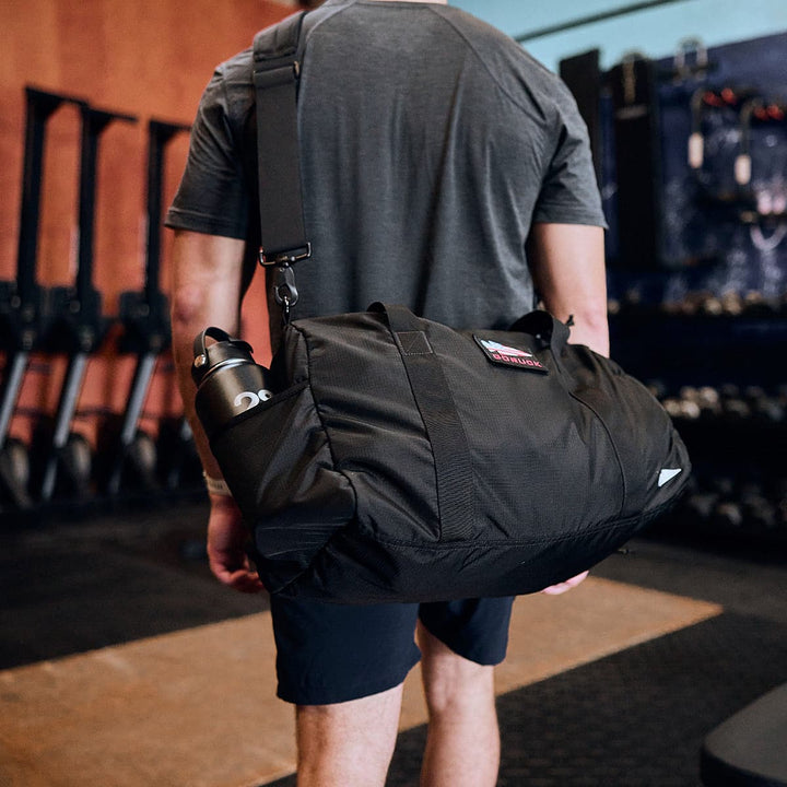 A person stands in the fitness area, carrying a GORUCK Gym Bag - Ripstop ROBIC®, with a water bottle nestled inside.