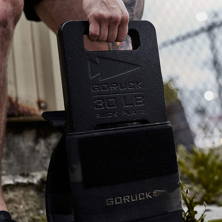 Effortlessly, a person lifts a 30 lb black Ruck Plates - LB by GORUCK outdoors, securely fastened in their Ruck Plate Carrier, with a sturdy fence providing the backdrop.