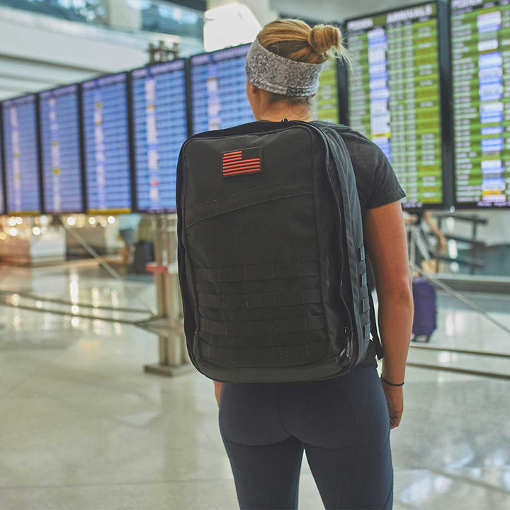 A traveler with a GORUCK GR2 - USA backpack is intently scanning the flight information screens at the airport, ensuring their carry-on compliant essentials are ready for the journey.