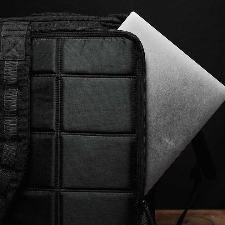 A silver laptop partially inserted into a black quilted GR2 - USA bag by GORUCK on a dark background, showcasing sleek style with 210D CORDURA® fabric for strength and carry-on compliant convenience.