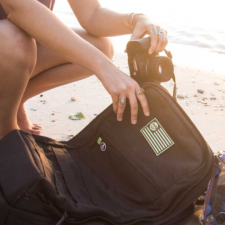 On a sandy beach, someone is carefully placing their camera into the GORUCK GR2 - USA, a sleek black backpack crafted from durable 210D CORDURA® fabric, ensuring it's carry-on compliant for their next adventure.