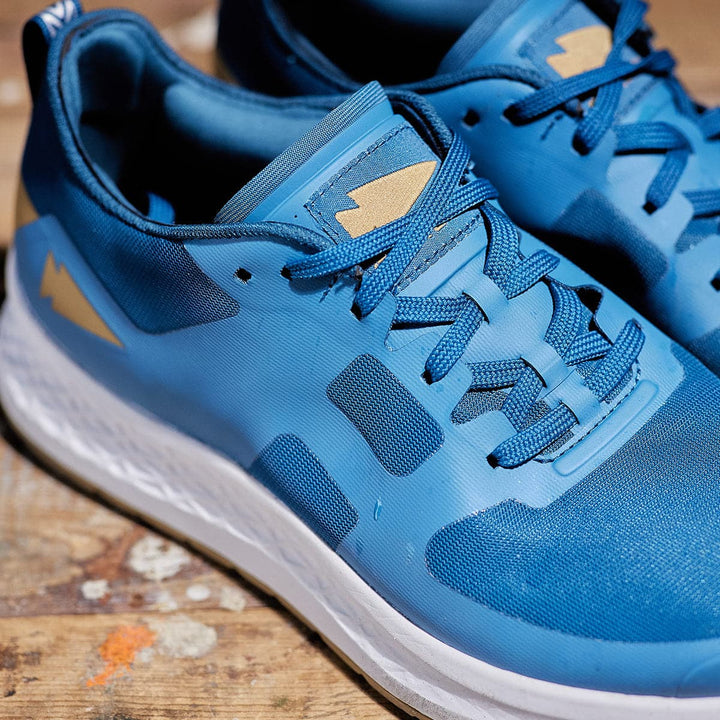 A close-up of GORUCK's Men's Rough Runner - Tidal Blue athletic shoes showcases their blue design with white soles and a subtle lightning bolt pattern, featuring Gradient Density EVA for enhanced high mileage performance. These laced-up Tidal Blue shoes are displayed on a paint-splattered wooden surface.