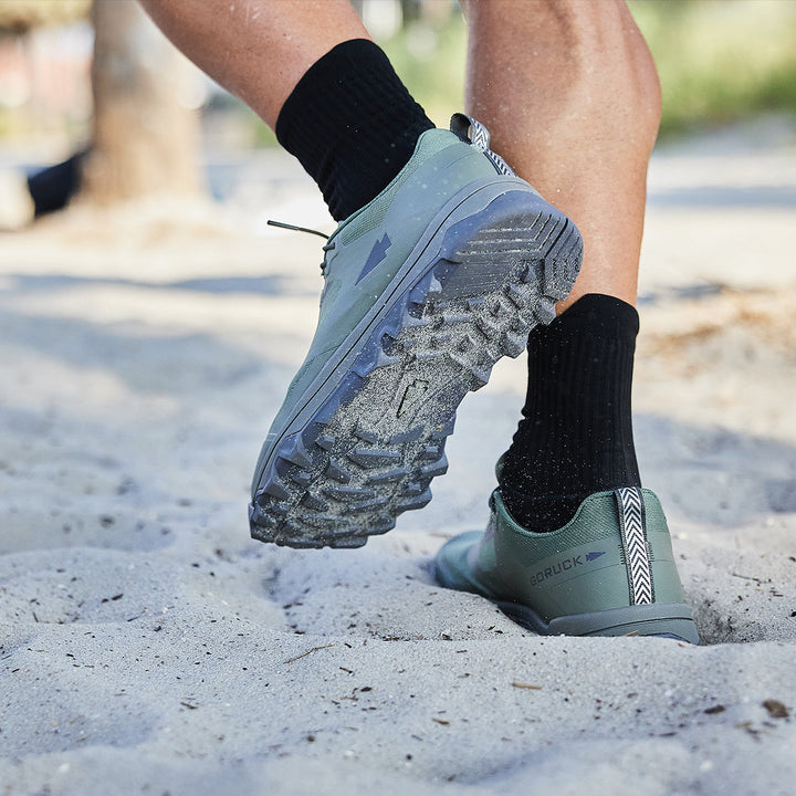 Close-up of a person running on sand, their feet clad in GORUCK's Mackall shoes in Earth and Forged Iron colors, paired with black socks, ideal for Special Forces training.