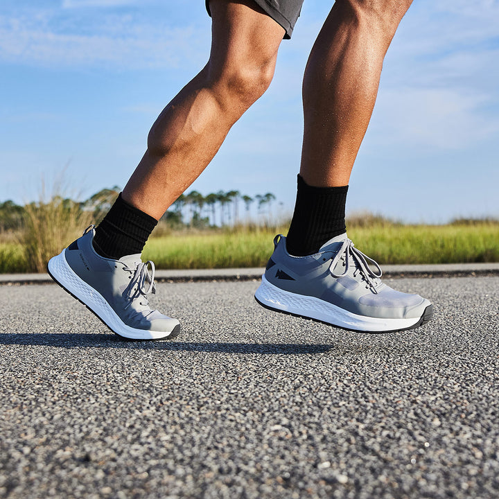 A person is running on a paved road, wearing GORUCK's Rough Runner - Speed Grey + White, ultra-supportive running shoes paired with black socks. The versatile road-to-trail outsole ensures stability, complementing the lush greenery in the background that adds a refreshing touch to their invigorating workout.