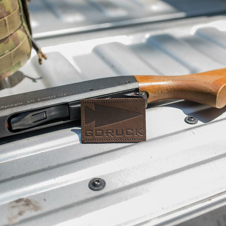 A shotgun rests on a metal surface, with a Patch - Leather GORUCK Spearhead by vendor-unknown placed on top.