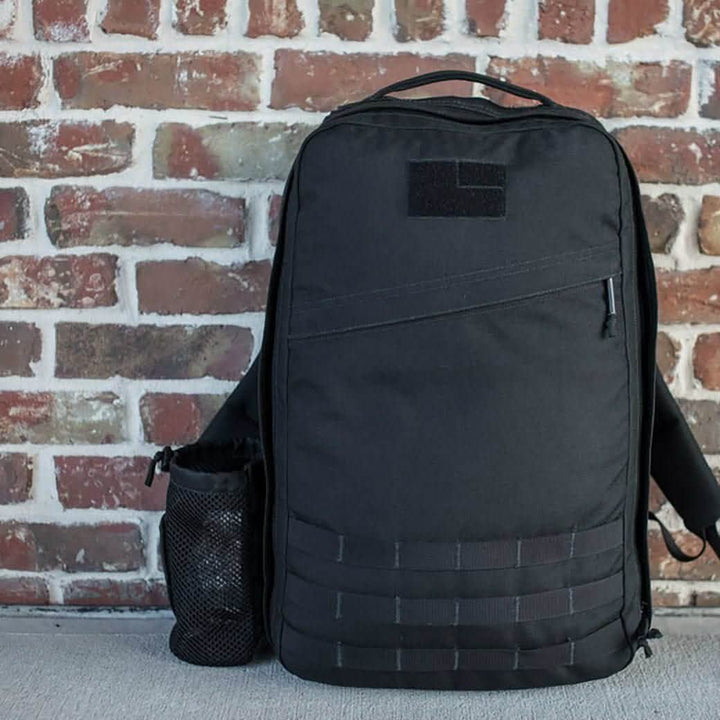 A black backpack with minimalist design from GORUCK is set against a rustic brick wall, showcasing its convenient portability. It features a small Velcro patch area and the Mesh Water Bottle Pocket, ideal for carrying your Nalgene water bottle, all placed on a light-colored surface.