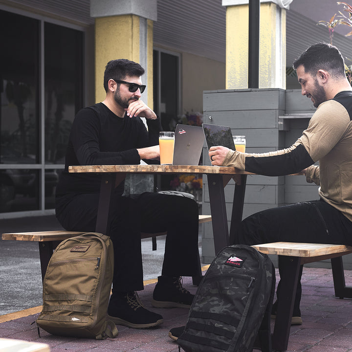 Two men at a wooden table work on laptops, while their GORUCK Bullet Ruck Laptop - 16L backpacks lie nearby, offering lumbar support and a bombproof compartment for safe laptop storage.