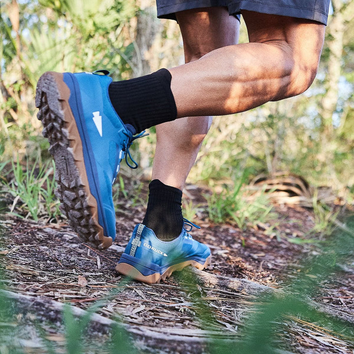 Wearing Mackall - Legion Blue + Gum trail running shoes from GORUCK, a person jogs on a forest path with an aggressive triple compound outsole.