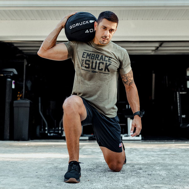 A man kneels on one leg wearing workout attire, holding a 40 lb Sand Medicine Ball by GORUCK on his shoulder and sporting an "Embrace The Suck" shirt. Ideal for boosting core stability during intense home gym sessions.