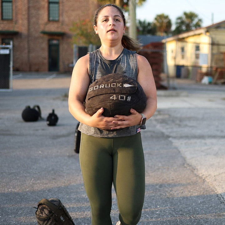 Amidst kettlebells and towering buildings, a person carries a Sand Medicine Ball by GORUCK outdoors, enhancing core stability as if in their own home gym.