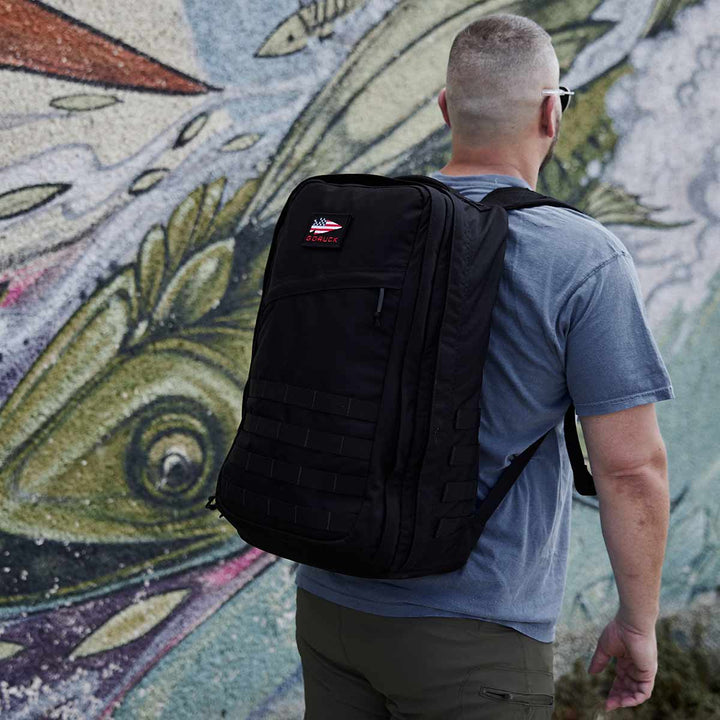 With a GR2 - USA black rucksack from GORUCK, a person stands in front of a colorful fish mural, possibly appreciating the art while relying on the Scars Lifetime Guarantee that ensures their bag accompanies them on all their journeys.