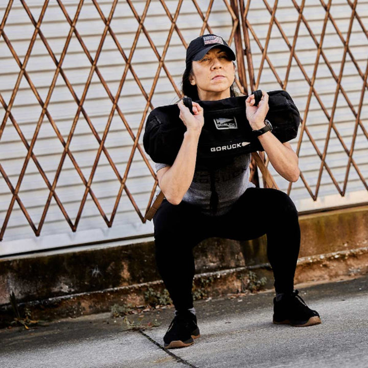 A person is squatting outdoors with the GORUCK Sandbag Kit (w/ Filler Bag) featuring heavy-duty handles, wearing a cap and athletic clothing. This durable gear promises top performance, backed by the Scars Lifetime Guarantee for challenging and reliable workout sessions.