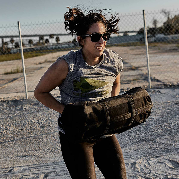 Wearing sunglasses and a gray tank top, a person runs outdoors gripping the Sandbag Kit's heavy-duty handles by GORUCK.