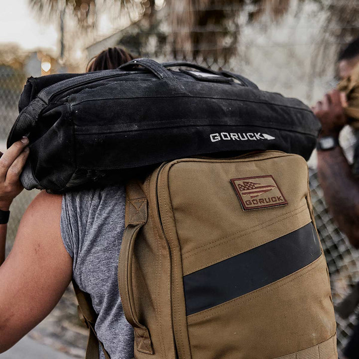 A person is carrying a large GORUCK Sandbag Kit with heavy-duty handles on their shoulders, while wearing a tan backpack outdoors.