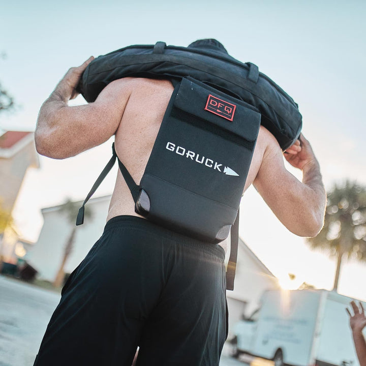 A man carries a heavy tire over his shoulders outdoors, wearing a black GORUCK Ruck Plate Carrier 3.0 made from durable ballistic nylon. This rugged accessory ensures comfort with ergonomic lumbar support, perfect for any adventure.