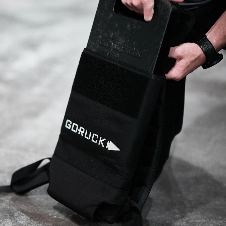 Inserting a weight plate into the Ruck Plate Carrier 3.0 by GORUCK, crafted from durable ballistic nylon, designed for the ultimate experience on a concrete surface.