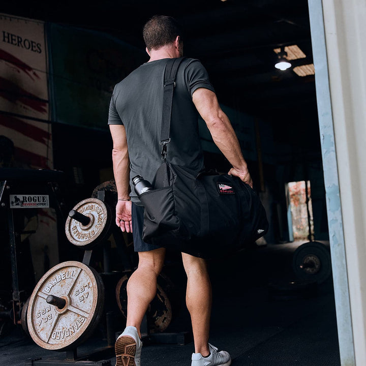 A man strides into the gym carrying a sleek GORUCK Gym Bag - Ripstop ROBIC®, crafted for durability. Weight sets and exercise equipment await him as he finds his rhythm, with the bag's dedicated shoe compartment keeping everything neatly organized.