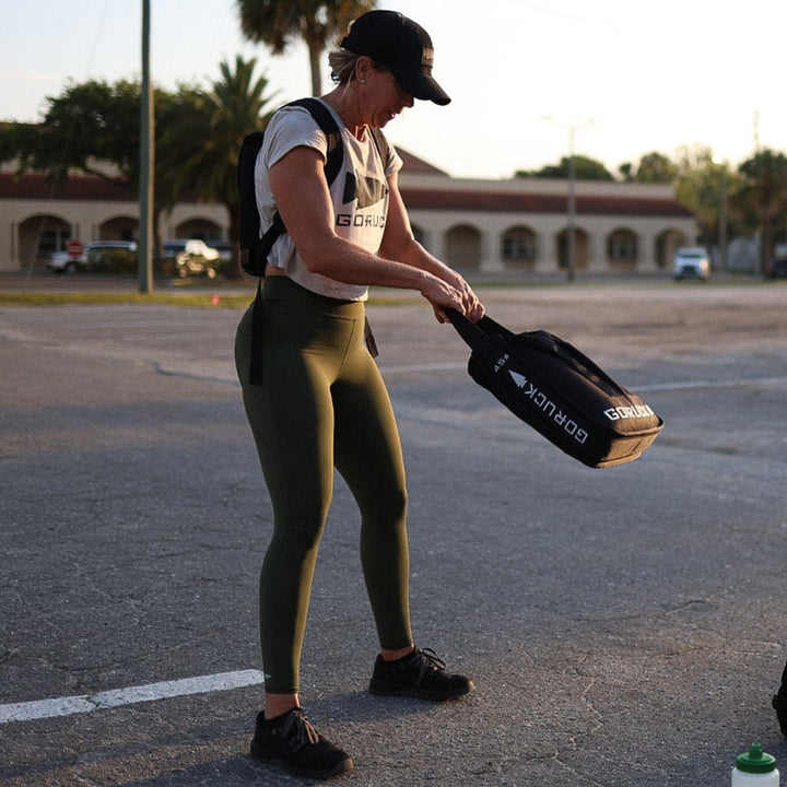 A person in activewear and a cap holds a Sand Jerry Can by GORUCK, constructed from Special Forces Grade materials, standing outdoors in a parking lot at sunrise.