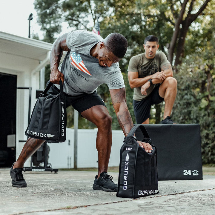 Two men are exercising outdoors; one lifts a GORUCK Sand Jerry Can, crafted with Special Forces Grade materials, while the other builds core strength sitting on a box.