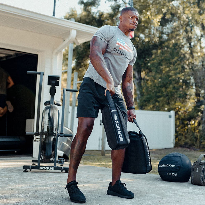 A man in a gray shirt and black shorts exercises outdoors using the Sand Jerry Can from GORUCK, crafted from special forces grade materials, to boost his core strength. Nearby, fitness equipment sets the scene for an intense workout.