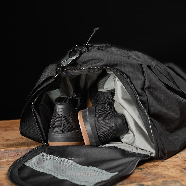 A Gym Bag - Ripstop ROBIC® by GORUCK, in black, rests on a wooden surface against a dark backdrop. Crafted from durable ripstop nylon, it reveals black sneakers nestled inside its shoe compartment.