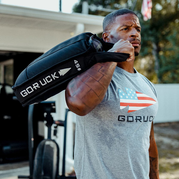 A man demonstrates his core strength by hefting a black GORUCK Sand Jerry Can, made from Special Forces Grade materials, on his shoulder. He's dressed in a gray GORUCK t-shirt featuring an American flag design.