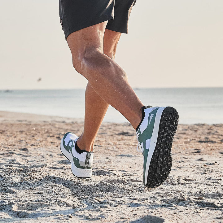 A Rough Runner jogs along the sandy beach in Men's Rough Runner - Seaspray by GORUCK, with the ocean visible in the background. The sun casts a warm glow, highlighting their muscular legs, while the Gradient Density EVA enhances their stride amidst nature's beauty.