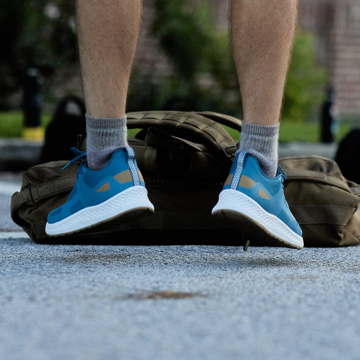 A person wearing GORUCK's Men's Rough Runner shoes in Tidal Blue and gray socks stands next to a large olive green bag on the sidewalk, prepared for high mileage. The focus is on their lower legs and feet, set against a blurred background of greenery and a brick wall.