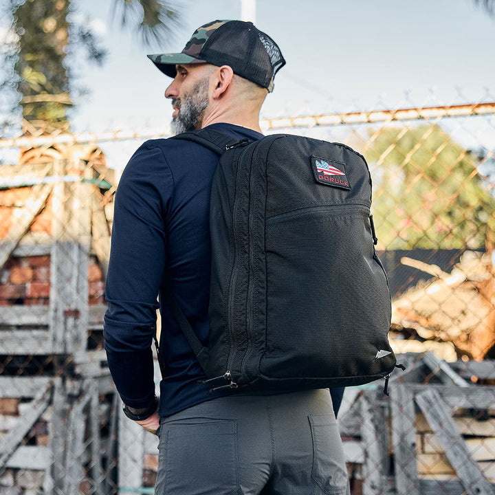 A man in a cap, carrying the GORUCK Bullet Ruck Double Compartment made of ripstop nylon, stands by a chain-link fence and wooden pallets.