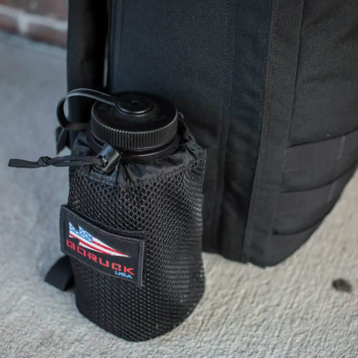 A black backpack rests on a textured cement surface, highlighting the GORUCK Mesh Water Bottle Pocket, which conveniently holds a Nalgene bottle. The pocket is adorned with an American flag patch and the GORUCK brand name, making it ideal for easy access while on-the-go.