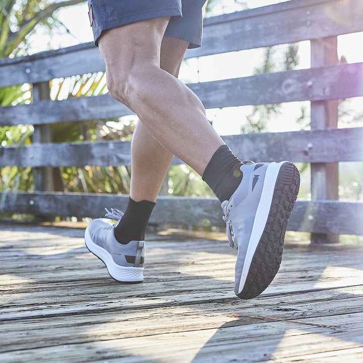 A person walks on a wooden deck wearing GORUCK's Rough Runner shoes in Speed Grey + White, featuring the GRADIENT DENSITY™ EVA MIDSOLE, along with black socks, enjoying the sunlight.