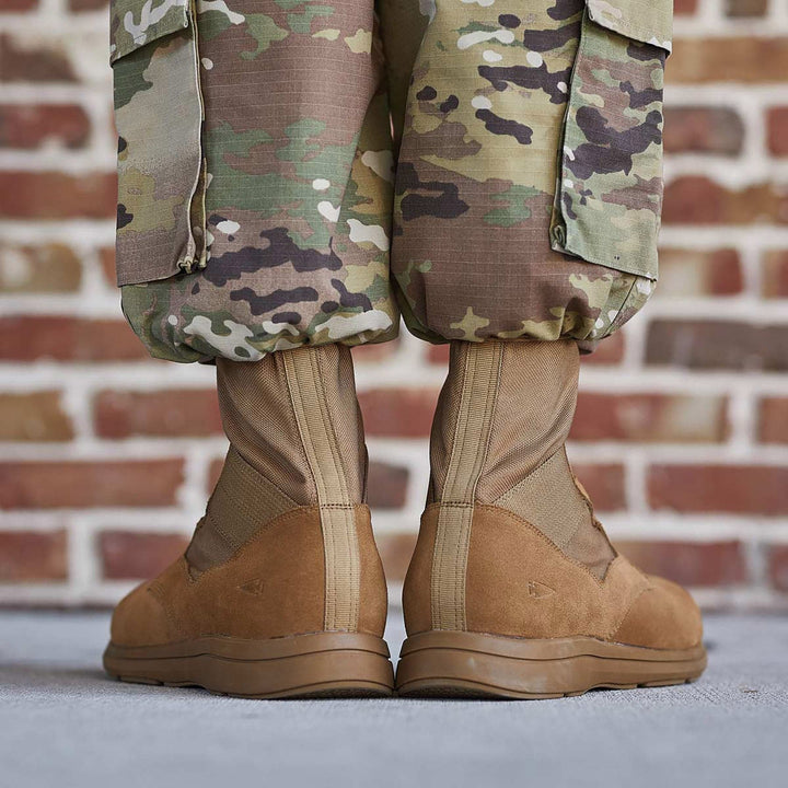 A person wearing camo pants and GORUCK's MACV-1 Hi-Speed high top boots, which are AR670-1 compliant, stands in front of a brick wall.