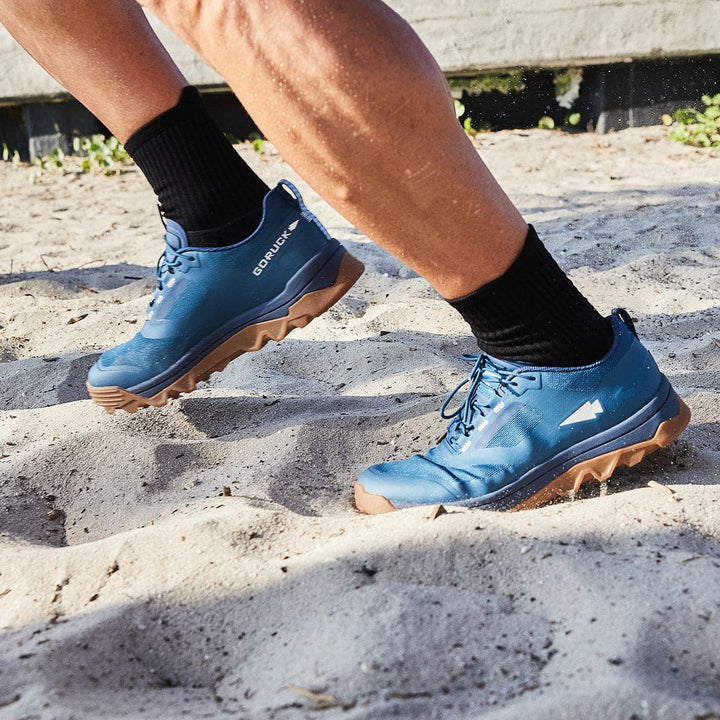 A person running on sandy ground wears the GORUCK Mackall in Legion Blue and Gum with black socks, featuring an aggressive triple compound outsole perfect for all-terrain rucking.