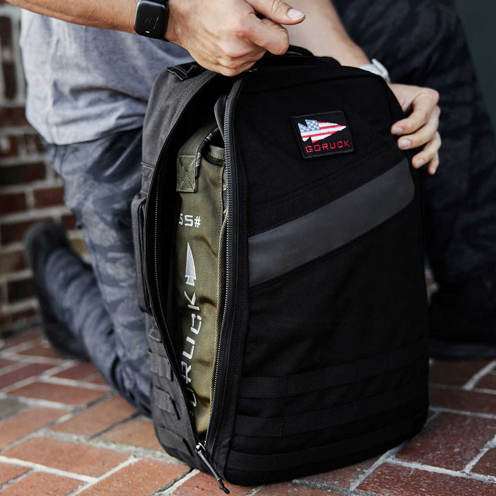 A person kneels, unveiling the packed contents inside a black Sand Jerry Can by GORUCK, crafted from Special Forces Grade materials.