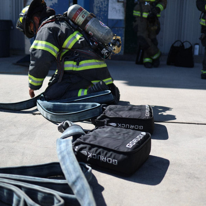 A firefighter kneels on the ground, sorting hoses near Sand Jerry Cans from GORUCK, crafted with Special Forces Grade materials, while another firefighter manages equipment in the background.