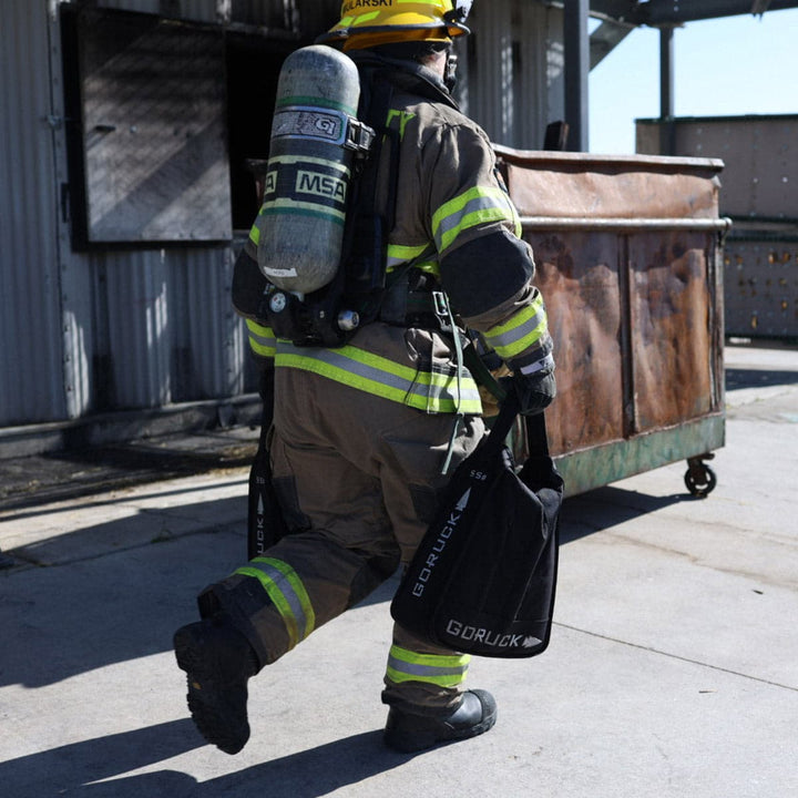 A firefighter in full gear demonstrates impressive core strength as they carry GORUCK Sand Jerry Cans during a rigorous training exercise near a metal dumpster.