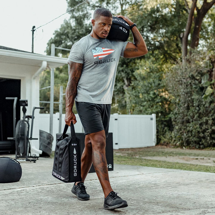 A person utilizes the Sand Jerry Can by GORUCK, made from Special Forces Grade materials, to exercise outdoors. They carry the weighted bags in both hands while wearing athletic attire near their home gym, concentrating on strengthening their core.