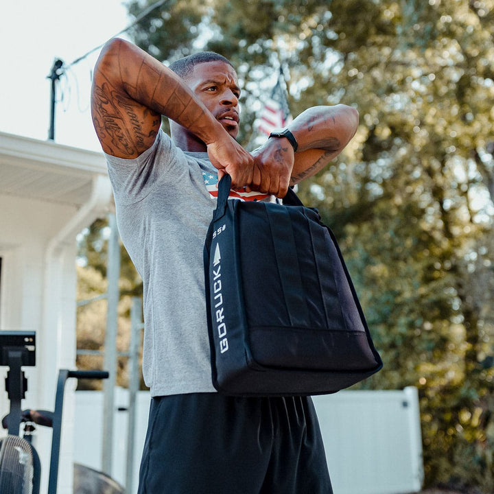 A person highlights their core strength by lifting a GORUCK Sand Jerry Can outdoors, dressed in a gray t-shirt and shorts. The Sand Jerry Can, constructed from Special Forces Grade materials, mirrors the rugged durability characteristic of its design.