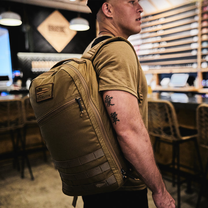 A person with a tan GORUCK Bullet Ruck Laptop - 16L and tattooed arms stands in a bar, showcasing stools and a menu board. The ruck's bombproof laptop compartment and lumbar support offer ultimate functionality in the lively setting.