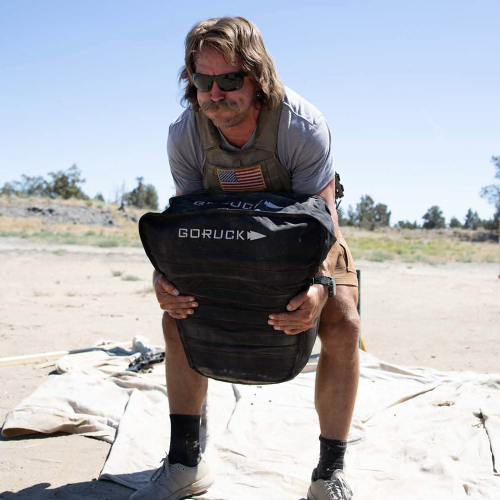 A person with long hair, wearing sunglasses and a military-style patch, hoists a weighted GORUCK Sand Tombstones backpack in an outdoor setting. Standing on sandy ground under a clear sky, they concentrate on their loaded carries workout, embodying the spirit of physical endurance.