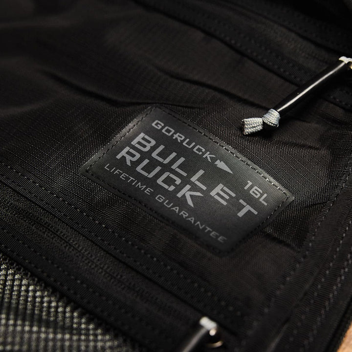 Close-up of a label on the GORUCK Bullet Ruck Double Compartment - Ripstop Nylon travel backpack displays "16L" and "Lifetime Guarantee" in black, emphasizing its suitability for adventures.