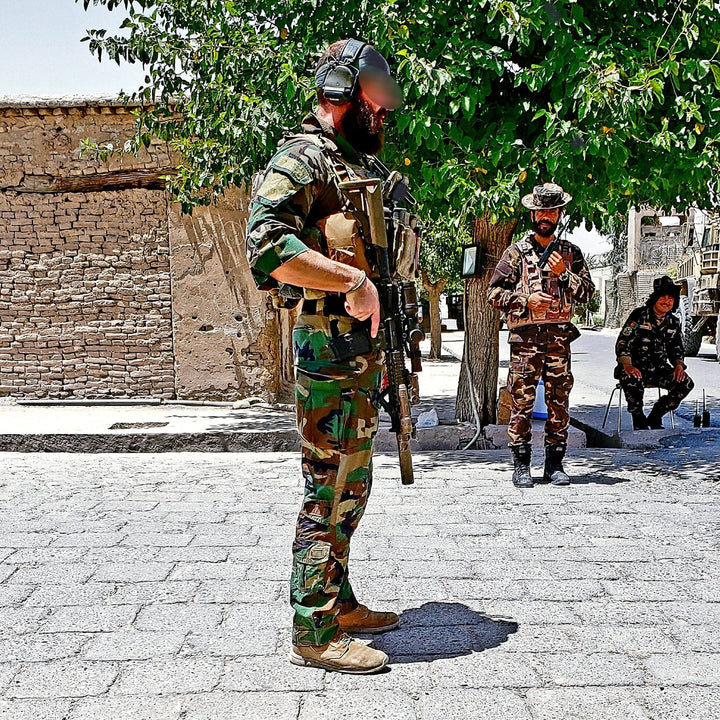 Armed soldiers in camouflage gear, wearing AR670-1 compliant and lightweight MACV-1 Hi-Speed - High Top rucking boots from GORUCK, stand on a sunlit street near a tree, with a brick building in the background.