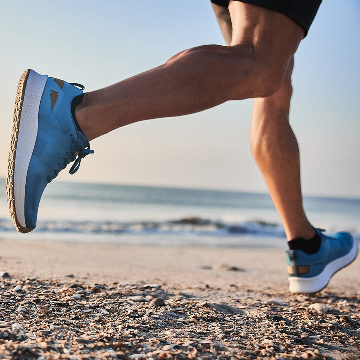 A GORUCK Men's Rough Runner - Tidal Blue sprints along the beach, showcasing sneakers with Gradient Density EVA, ideal for tackling high mileage. The sandy shore stretches out in the foreground while the ocean sparkles under a clear sky in the background.