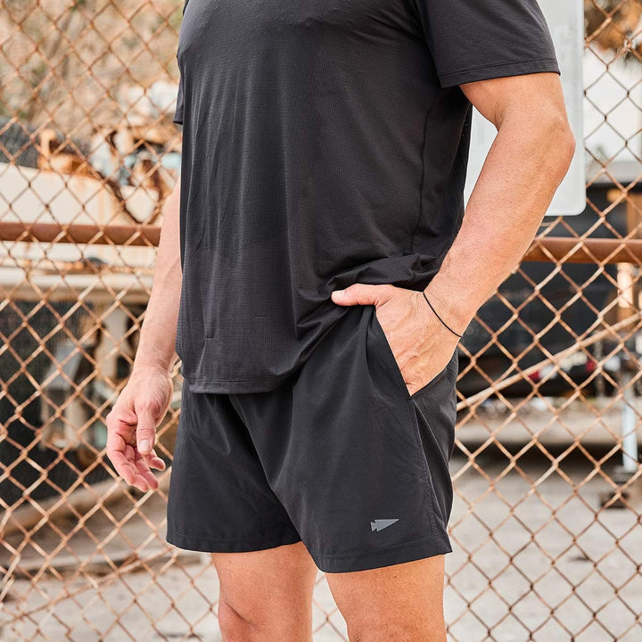 A person in a black athletic outfit stands with one hand in their pocket in front of a chain-link fence, wearing GORUCK's Men’s Training Shorts made from ToughStretch fabric, which perfectly complement the T-shirt. The blurred industrial elements in the background add an edgy vibe.